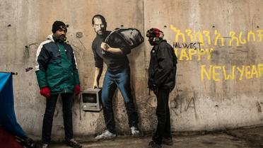 Des migrants devant une oeuvre de l'artiste britannique Bansky photographiée le 12 décembre 2015 et située à l'entrée de la "Jungle" de Calais, représente Steve Jobs (Apple), qui porte un baluchon et un vieil ordinateur [PHILIPPE HUGUEN / AFP]
