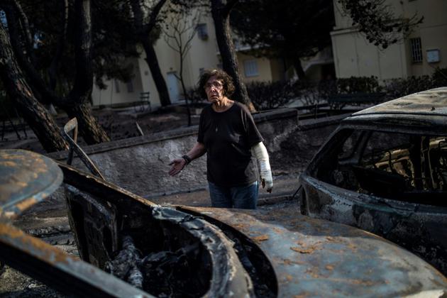 Une femme, le bras plâtré, devant des voitures brûlées après l'incendie meurtrier qui a dévasté Mati, le 24 juillet 2018 près d'Athènes [ANGELOS TZORTZINIS / AFP]