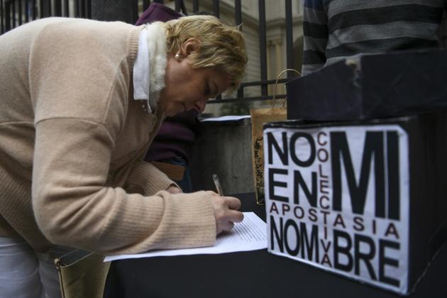 Une femme paraphe un document renonçant à son baptême à côté d'une pancarte disant "Pas en mon nom. Apostasie collective", devant le siège de la conférence épiscopale argentine, le 24 août 2018 [EITAN ABRAMOVICH / AFP]